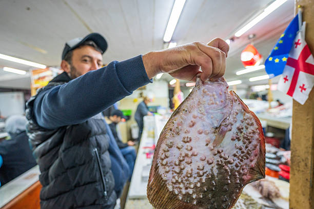 Batumi, Georgia - October 24, 2024: At the vibrant fish market in Batumi, Georgia, a local vendor proudly displays a freshly caught flatfish to potential customers. This bustling coastal city on the Black Sea is known for its rich fishing traditions and lively local commerce. The photo captures not only the catch of the day but also the cultural essence of Georgia’s seafaring communities, as seen in the background details like the national and EU flags and Georgian signage