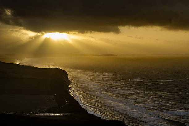 Sunlight streams through breaks in dark clouds, casting beams over a rugged sea cliff and ocean. The coastline is steep, with waves gently lapping the shore. The sky transitions from dark gray to a bright yellow where the sun illuminates the scene. The contrast between the darkened cliff and the illuminated ocean creates a dramatic and serene landscape.