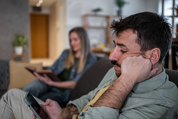 istock Bored man browsing smartphone while woman reading book on sofa 2215098882