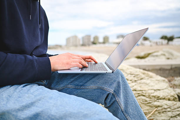 Young man working on his laptop while sitting on a rock by the sea