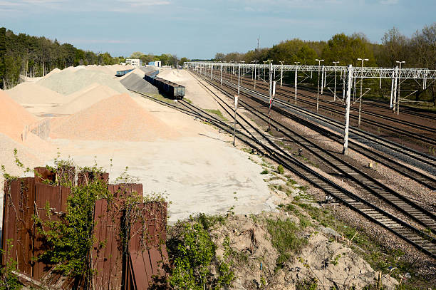Wide-angle view of a railway cargo terminal with multiple freight wagons loaded with aggregates such as gravel, sand, and crushed stone. Neatly organized piles of construction materials lie adjacent to the railway tracks, with an overhead catenary system indicating electrified lines. Surrounded by trees and a clear sky, the scene illustrates industrial infrastructure, rail-based logistics, and material supply chains for construction and development industries.