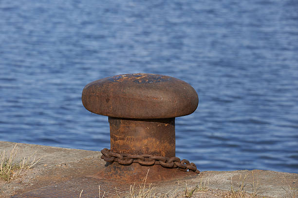 A rusted bollard, adorned with securing chains, is positioned along an aged dock while the tranquil water stretches beyond. The scene evokes maritime tradition, industrial wear, and timeless oceanic essence.