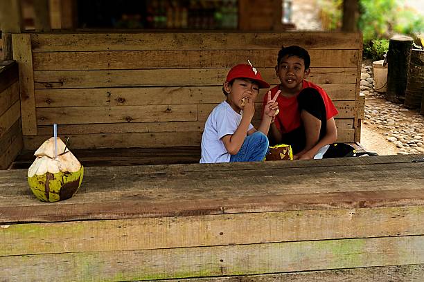 Two boys enjoy snacks and fresh coconuts while relaxing at a traditional small wooden stall in a peaceful rural setting