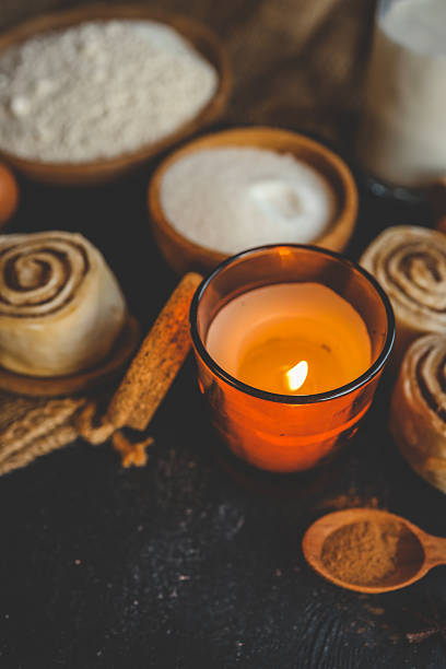 A cozy baking scene with cinnamon buns, a lit candle, flour, sugar, eggs, and milk on a dark surface, perfect for warm, rustic kitchen themes
