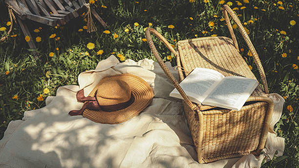 Woven picnic basket with an open book on top, resting on a cream blanket surrounded by blooming dandelions and a straw sunhat on a sunny spring day