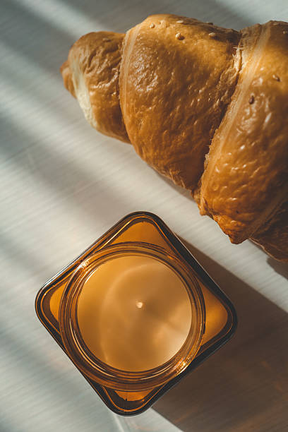 Golden croissant and small glass jar candle placed on white tray, lit by striped morning sunlight through window blinds, creating warm shadows and a cozy, rustic feel
