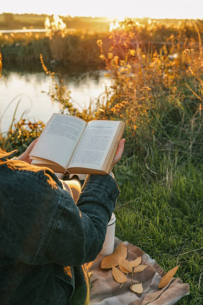 istock Girl by the river reading a book, beautiful sunset 2215162419