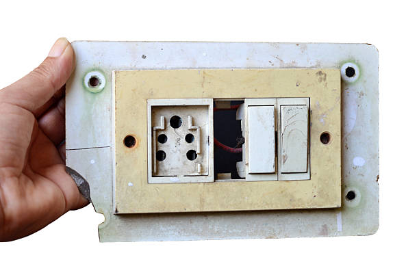 Cut out of rusty rustic run down weathered distressed damaged run down broken messy dirty electrical board plate, three pin socket, two switches, one missing part making a hole held in the hand of an electrician or repairman isolated over white background
