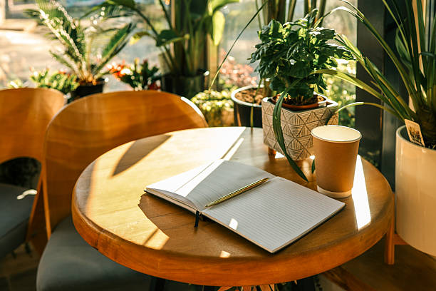 Open notebook with golden pen and takeaway coffee cup on wooden table in sunlit cafe with green houseplants and cozy modern interior