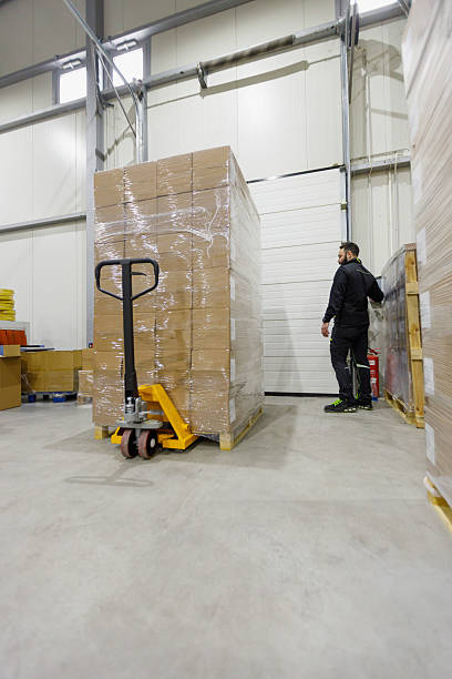istock Man by stack of cardboard boxes on pallet jack in warehouse, blue collar at workplace 2215164567