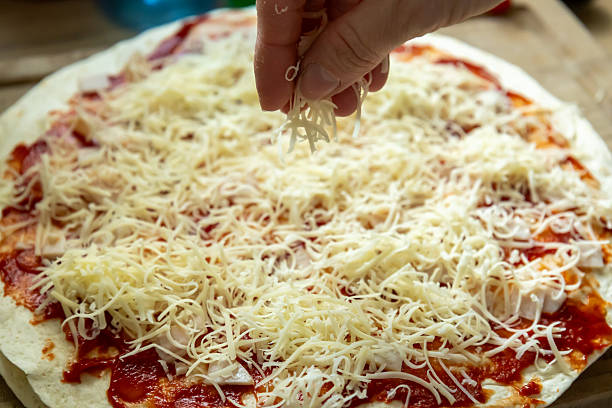 A hand is adding grated cheese over a tortilla spread with tomato sauce and diced ham. This step highlights the final touch in preparing a delicious homemade wrap.