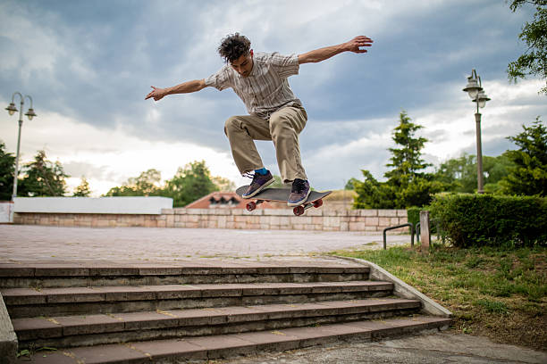Action shot of a young, stylish Gen Z man wearing casual pants and a loose shirt, performing a jump on his skateboard over a set of stairs in a public park. Captured mid-air, this dynamic scene showcases urban energy, youth lifestyle, and skateboarding culture.