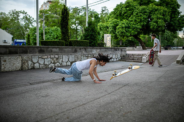 A young Gen Z man skateboarding with his friend in a public park falls off his board while attempting to jump over a stone wall. The scene captures a real moment of skateboarding practice, highlighting that failure and falls are part of learning new tricks and improving skills