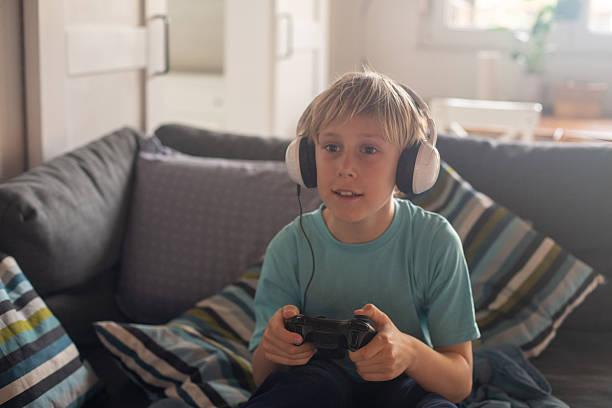 Smiling freckled boy with headphones enjoying video game on console while sitting on sofa at home.