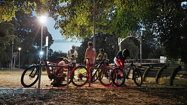 Group cyclists examining and preparing their mountain bikes near a parking area at dawn under streetlights
