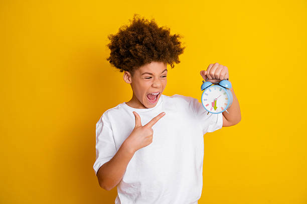 istock Excited boy pointing at retro clock in vibrant yellow background conceptualizing time with energetic expression 2215194528