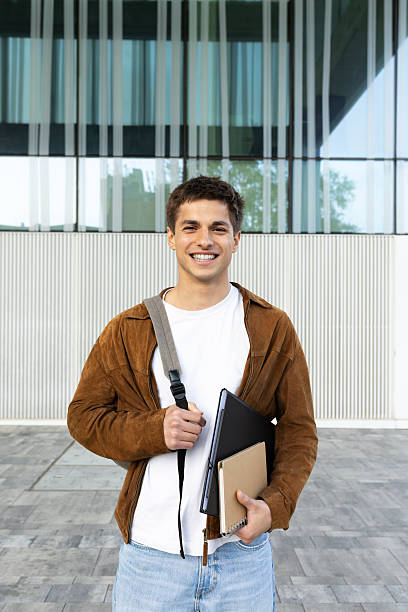 istock Smiling university student holding laptop and notebooks in front of modern building 2215204672