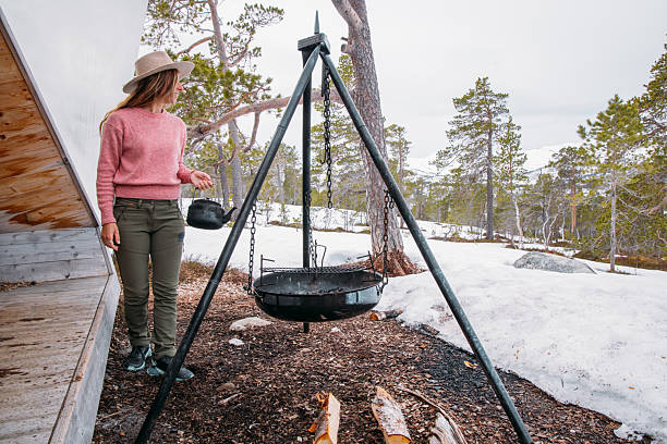 Woman in pink sweater and hat setting up a black kettle on a campfire tripod. Outdoor winter camping scene with snowy pine forest. Ideal for adventure and travel themes.