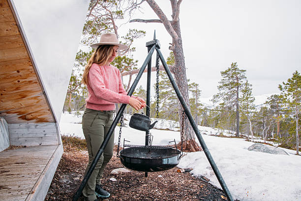 Woman in pink sweater and hat setting up a black kettle on a campfire tripod. Outdoor winter camping scene with snowy pine forest. Ideal for adventure and travel themes.