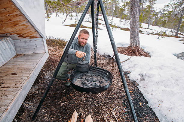 Man in winter forest setting heating coffee on grill, surrounded by snow. Seeking warmth and adventure during a camping trip. Rustic outdoor cooking experience.