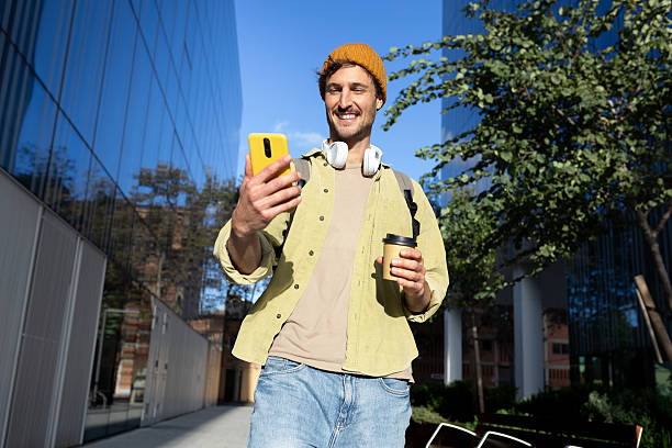 Smiling young man using smartphone and drinking coffee outdoors