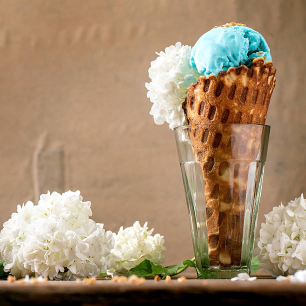 Turquoise ice cream with white flowers in waffle cone, standing in transparent glasses on wooden table. Trendy homemade summer dessert. Copy space
