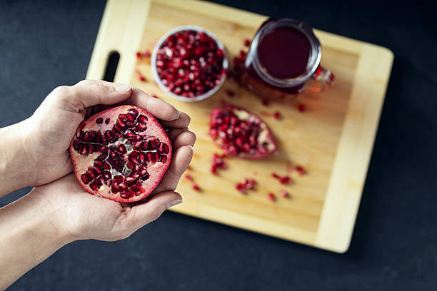 istock Hands holding cutted pomegranate and a ceramic bowl full of pomegranate seeds, cutted pomegranate and a glass of pomegranate juice on the background lie on the wooden cutting board. View from the top 2215216548