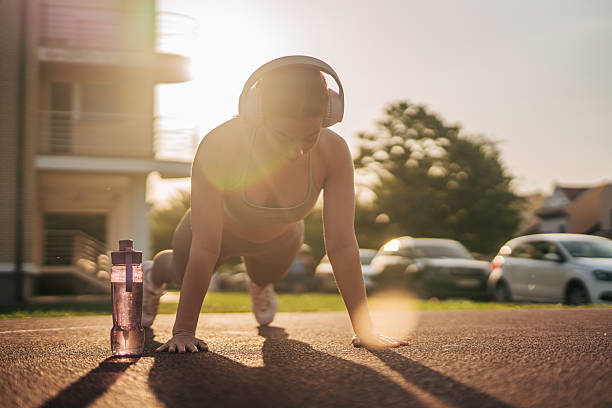 Young sportswoman listening to music while performing push-ups outdoors on a track and field, enjoying the vibrant colors of a beautiful sunset during the golden hour