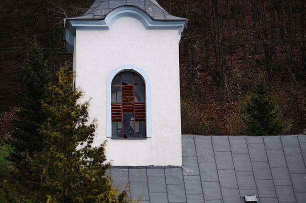 A white church tower with an arched window and light blue trim stands against a forested hillside. The wooden shutters and curved architectural details give the structure a quaint, historic charm amid the natural setting.