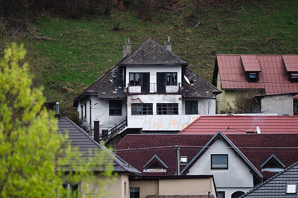 An old, abandoned house with broken windows and a deteriorating white facade stands on a grassy hillside, partially hidden behind the rooftops of well-maintained homes. The scene contrasts neglect with modern residential upkeep.