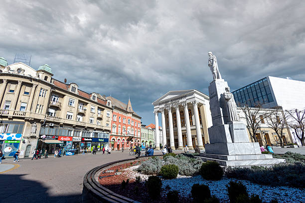 Subotica, Serbia - March 8, 2025: Wide-angle view of a European city square featuring classical architecture, a marble statue, and dramatic clouds. Pedestrians stroll by retail shops and historic buildings.