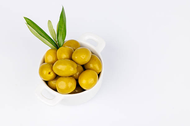 White bowl full of organic green olives placed next to an olive twig, on white background.