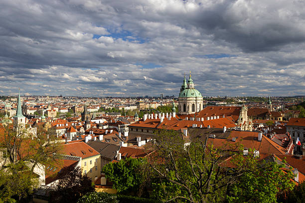 Panoramic view of Prague's red roofs and St. Nicholas Church under a dramatic cloudy sky, showcasing the city's architectural beauty