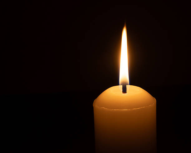 istock A single burning candle flame or light glowing on a big white candle isolated on black or dark background on table in church for Christmas, funeral or memorial service 2215258795