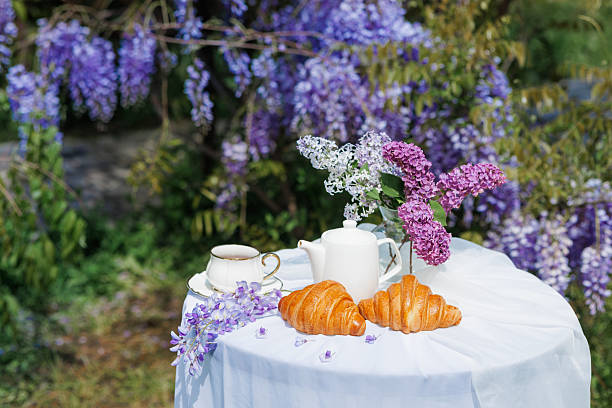 Spring breakfast scene with croissants, tea set, and lilac flowers on a white table in blooming wisteria garden.