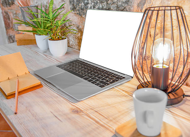 Cozy home office setup featuring a laptop with a blank white screen on a wooden desk. The desk is decorated with potted plants, a notebook, a ceramic cup, and a modern industrial-style lamp. Stone wall in the background adds a rustic touch. Ideal environment for remote work, freelancing, or study.