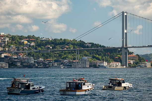 istock Fishing boats on the Bosphorus with large ships passing under the Bosphorus Bridge in Istanbul, Turkey on a bright day with scenic clouds, hills, and a Turkish flag in the background 2215269703