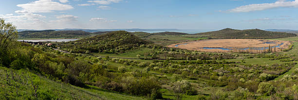 A sweeping panoramic view of the Tihany Peninsula reveals lush spring greenery, rolling hills, and the golden reed beds of the wetlands. The distant blue waters of Lake Balaton glisten under a bright sky dotted with soft clouds.