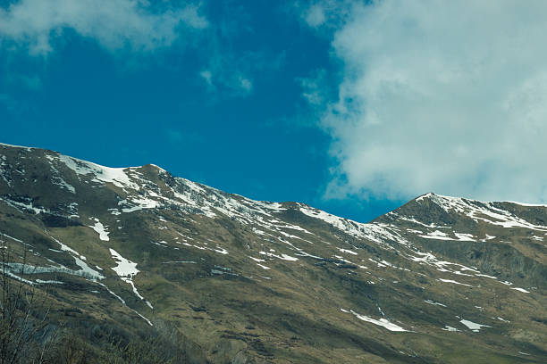A powerful view of the Caucasus Mountains in Georgia, where snow still blankets the upper peaks while the lower hills transition into spring. The stark contrast between the white snow and dark storm clouds above creates a dramatic and atmospheric scene. A few trees and patches of green show signs of renewal in this rugged yet beautiful landscape.