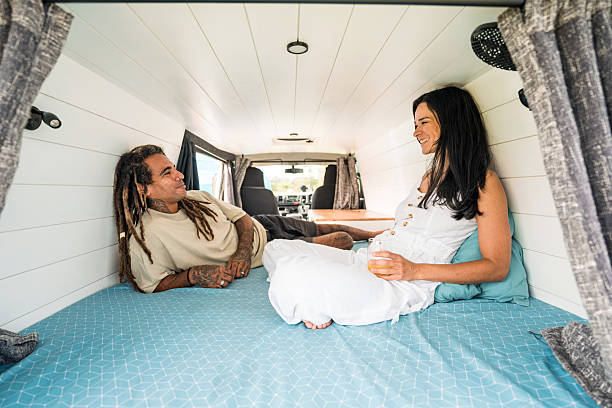 A multiracial mid-adult male with dreadlocks and a Hispanic female in casual attire relax on a bed inside a cozy camper van, bathed in soft natural light, sharing a joyful camping moment.
