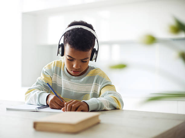 istock Dark-skinned seven-year-old boy doing homework with headphones. Writing in notebook in modern kitchen. 2215303787