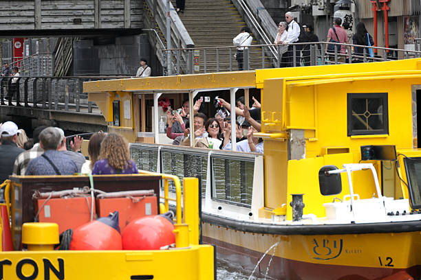 Osaka, Japan - April 12, 2025: Passengers in bright yellow tour boats wave to each other in the Dotonbori Canal. The Tazaemon Bridge with a pedestrian walkway spans the river. Spring afternoon in the Chuo Ward.