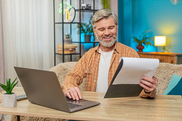 istock Mature man sitting on table with documents and laptop, smiling from successful work at home office 2215311626