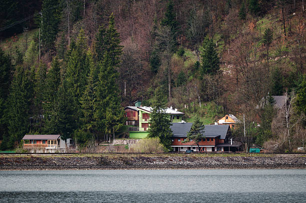A row of colorful houses sits at the base of a densely forested hillside, facing a calm lake. The lush greenery and peaceful water create a serene and secluded atmosphere in this scenic mountain location.