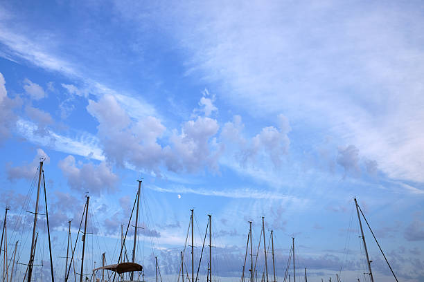 Tall sailboat masts rise into a dramatic blue sky streaked with soft clouds at a peaceful marina, creating a sense of calm and coastal rhythm. The minimalist composition emphasizes space, serenity, and the quiet elegance of life by the sea.