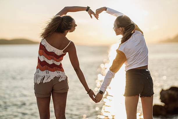 A happy female friends spending time on the beach, making a heart shape with their hands and enjoying in the sunset.