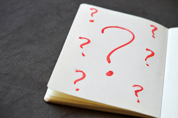 Several small red colored Question mark signs or symbols(one big standing out), in red sketch pen over white coloured paper of open vintage retro style notepad making white background as copy space placed over black textured blackboard or slate. There is ample copy space and no people.