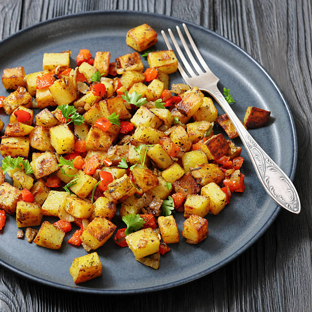 golden fried potatoes cubes with red bell pepper on a dark grey plate with fork on a black wooden table,  close-up