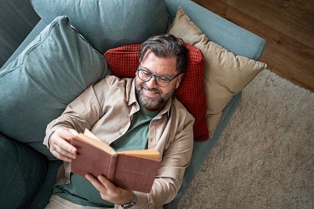 istock Smiling mature man relaxing on sofa reading a book 2215393356