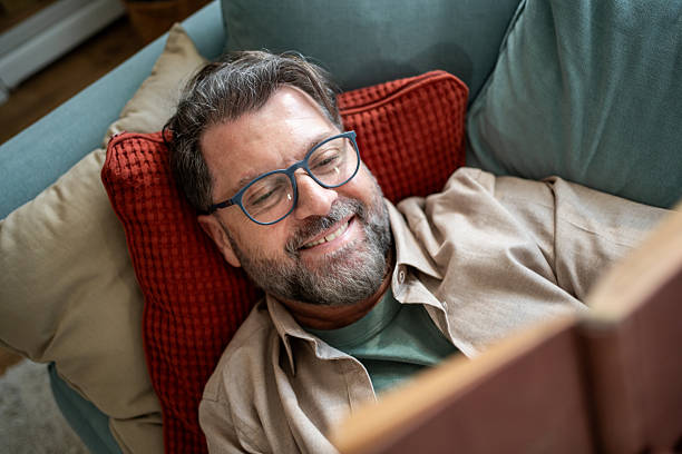 istock Mature man lying on sofa reading a book and smiling 2215393373